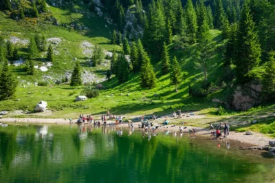 Obala Krnskega jezera ob katerem so taborniki, ki so tam na izletu. V ozadju zelene smreke, skale in del hriba.